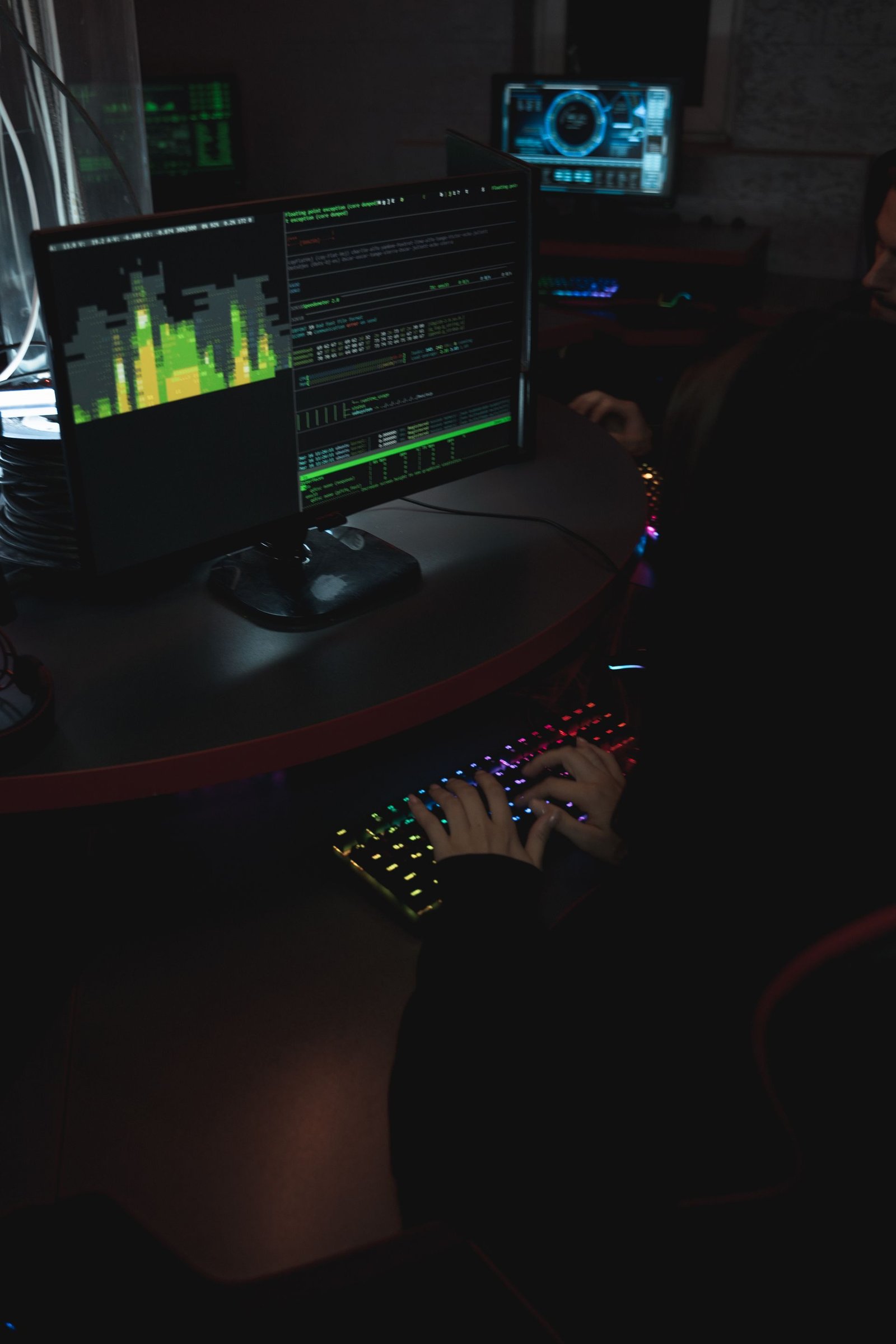 Hooded figure at a dark workstation with terminal monitors and a backlit keyboard